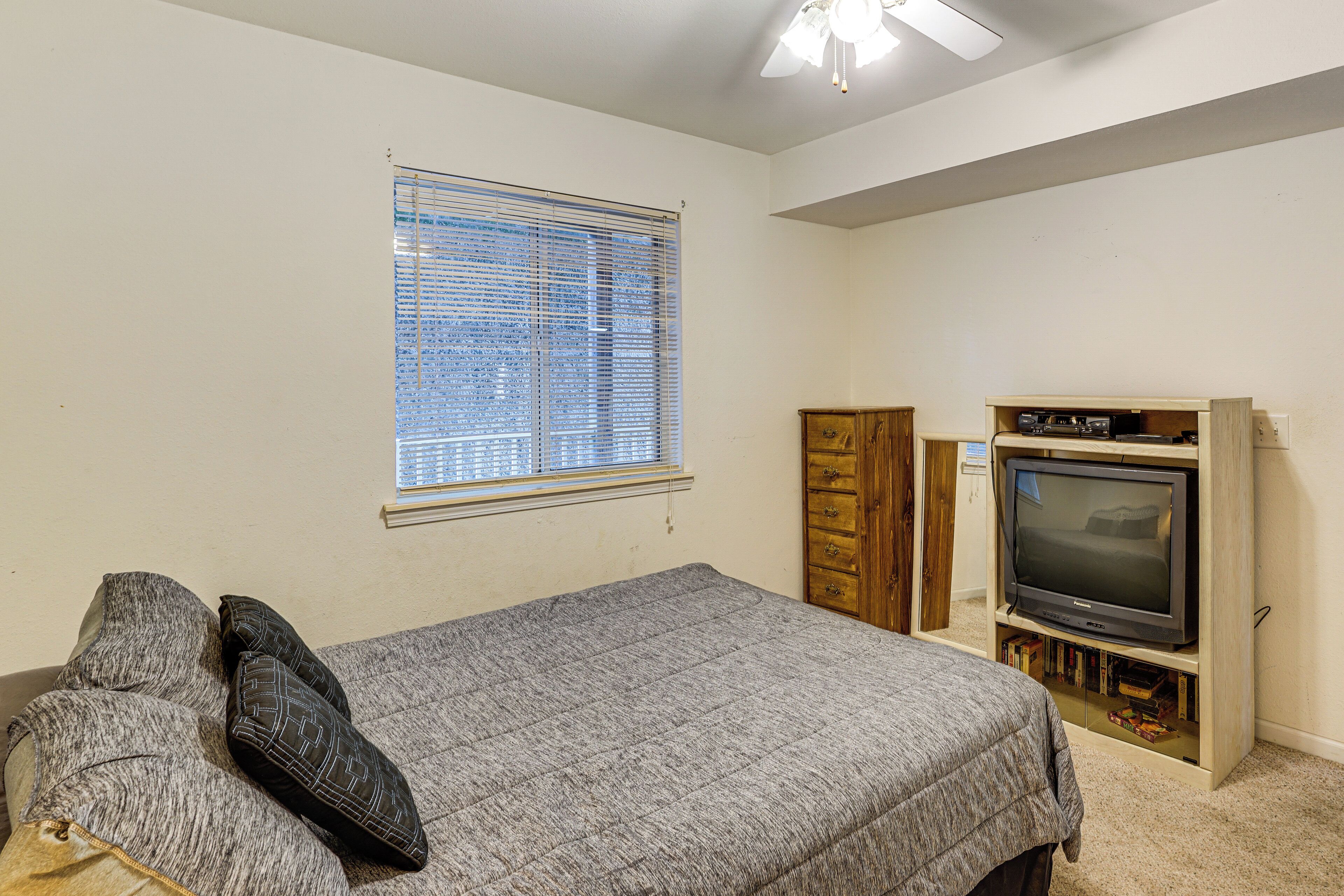 Une chambre à coucher avec un lit, une fenêtre avec des stores, un téléviseur posé sur un meuble et une armoire en bois.