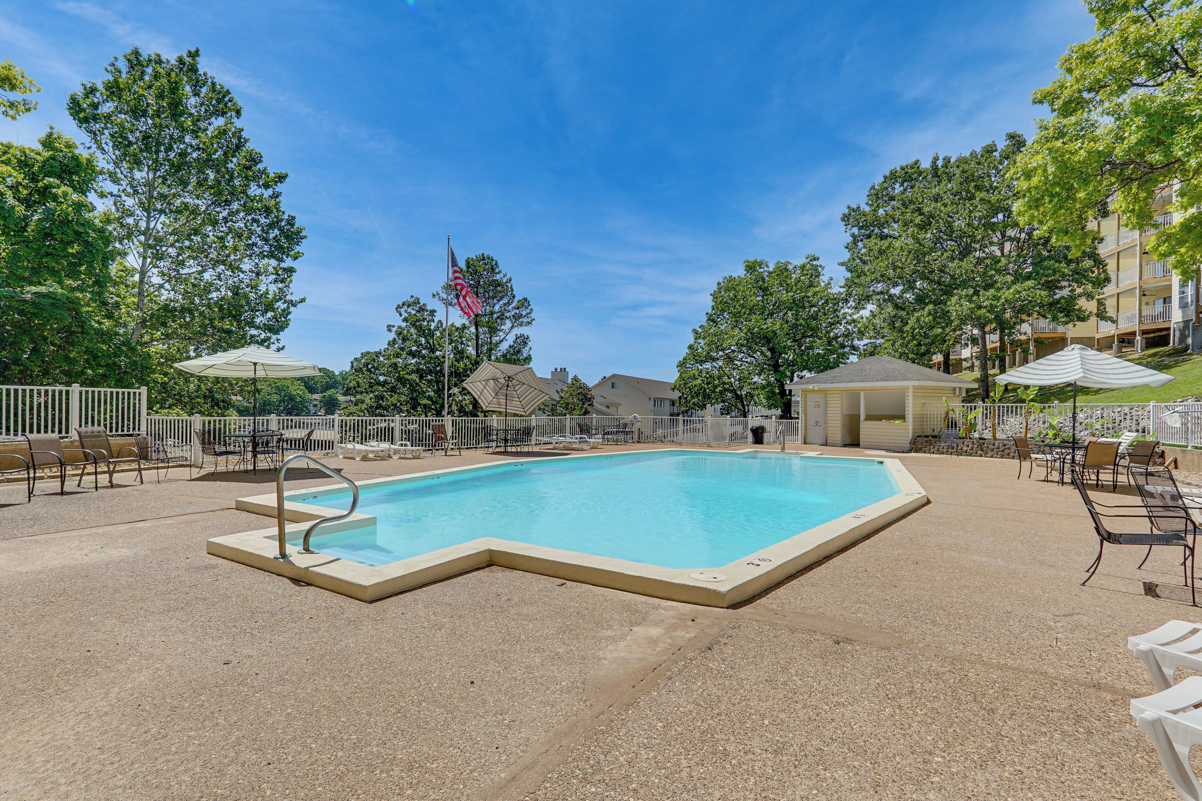 Une piscine rectangulaire avec un escalier en métal, entourée de chaises et de parasols, avec des arbres et un bâtiment en arrière-plan.