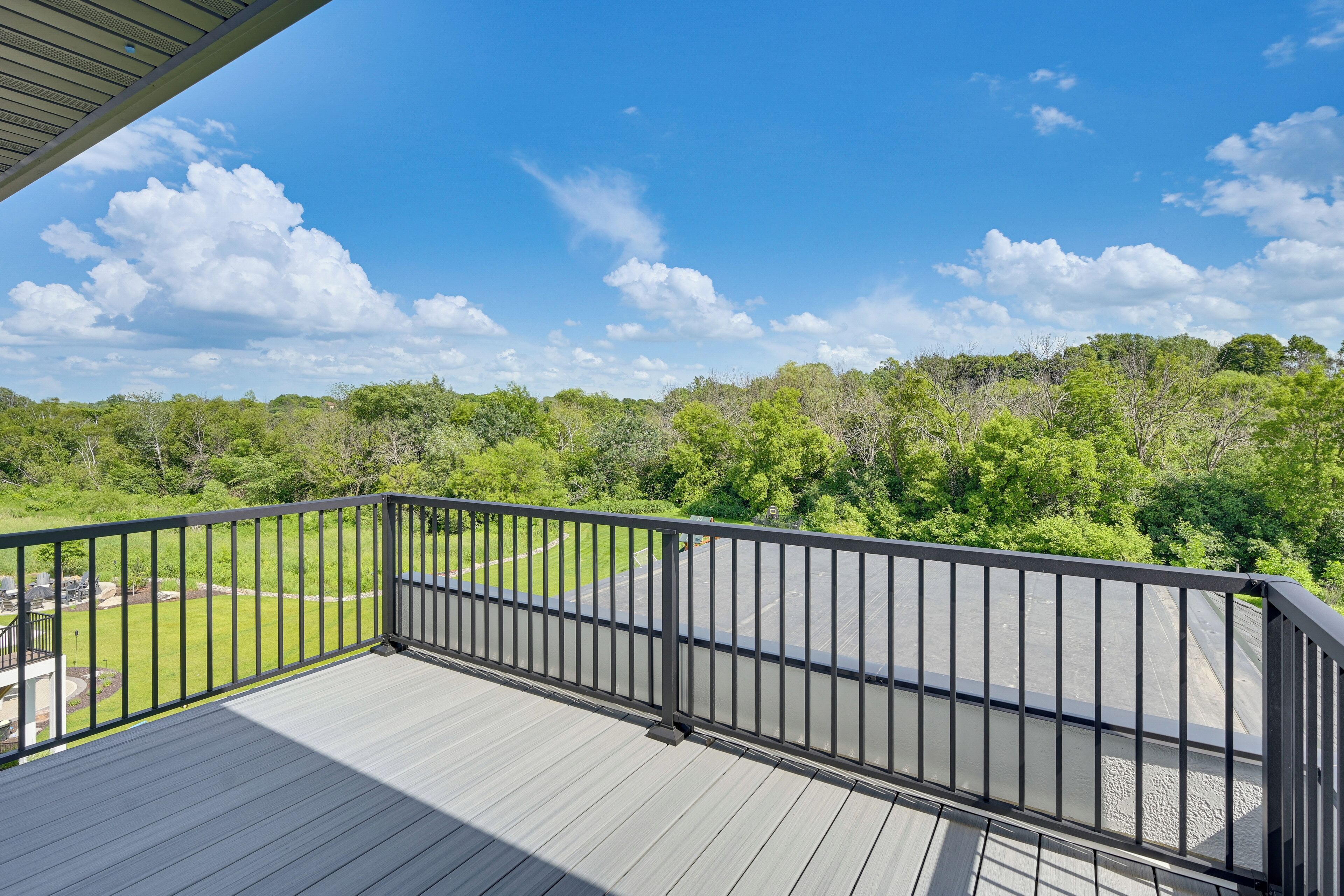 A deck with a black railing overlooking a green landscape and a parking area.