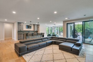 A modern living room with a sectional sofa, a kitchen area with a bar, and a large sliding glass door leading to an outdoor area. - Luxe Plymouth Group Getaway w/ Office & Foosball (Plymouth)