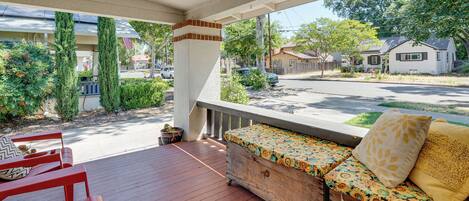 A covered porch with a wooden bench and red chairs, overlooking a residential street.