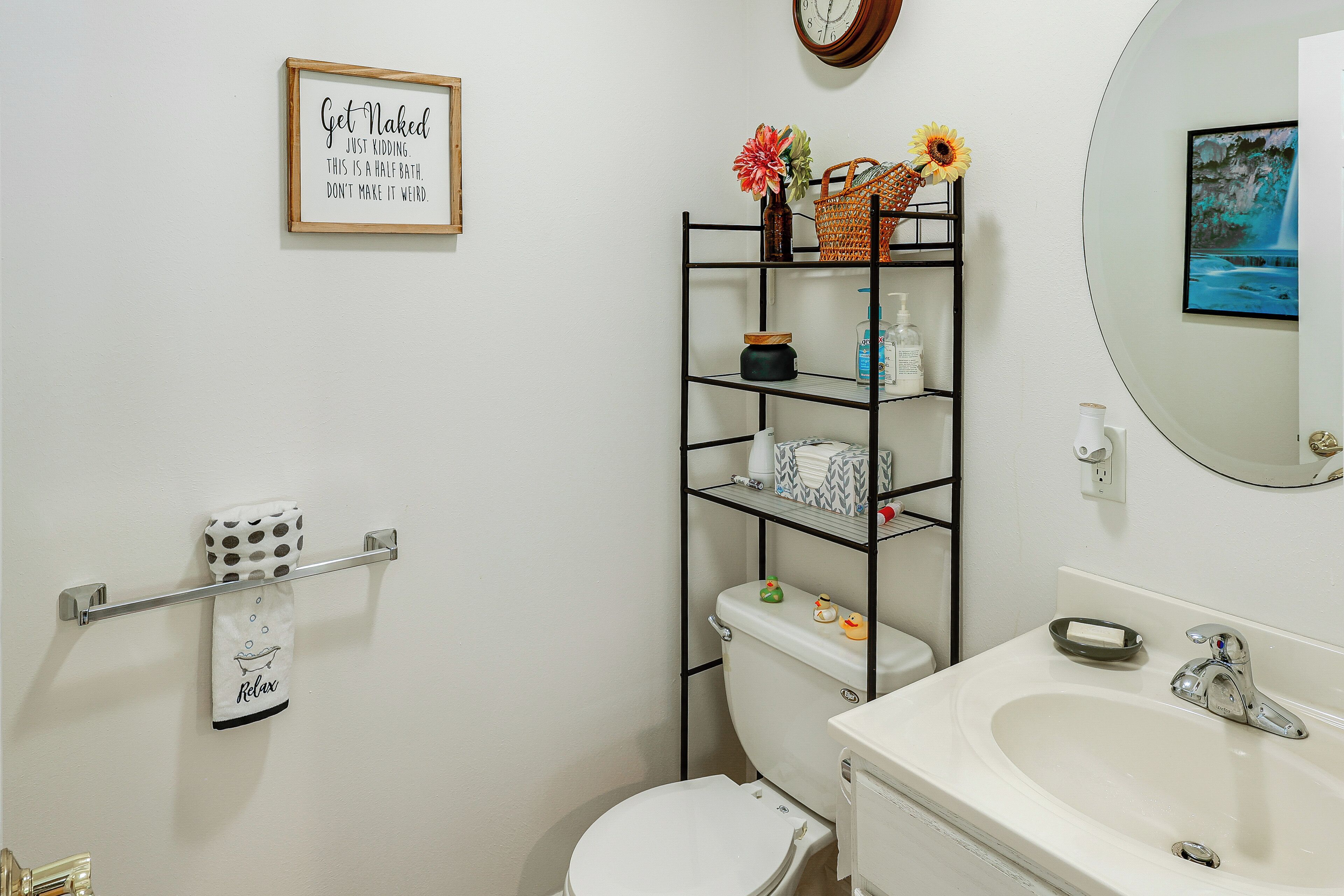 Une salle de bain comprenant des toilettes, un lavabo, une étagère avec des articles de toilette, un porte-serviettes et une citation encadrée au mur.