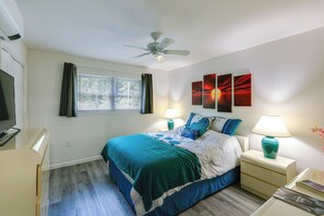 A bedroom with a bed, two bedside tables with lamps, a ceiling fan, a window with curtains, and artwork on the wall.