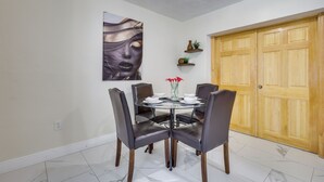 A dining area with a round table, four chairs, a vase with red flowers, a wall-mounted painting, and a wooden door. - Close to Hard Rock Stadium! Group Getaway w/ Yard (Miami Gardens)