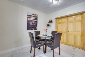 A dining area with a round table, four chairs, a vase with red flowers, a wall-mounted painting, and a wooden door.