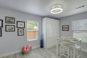 Una habitación con una mesa de comedor, sillas, un armario blanco, cuadros en la pared y una ventana con persianas.