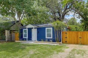 A small blue house with a front porch, a wooden fence, and a tree in the background.