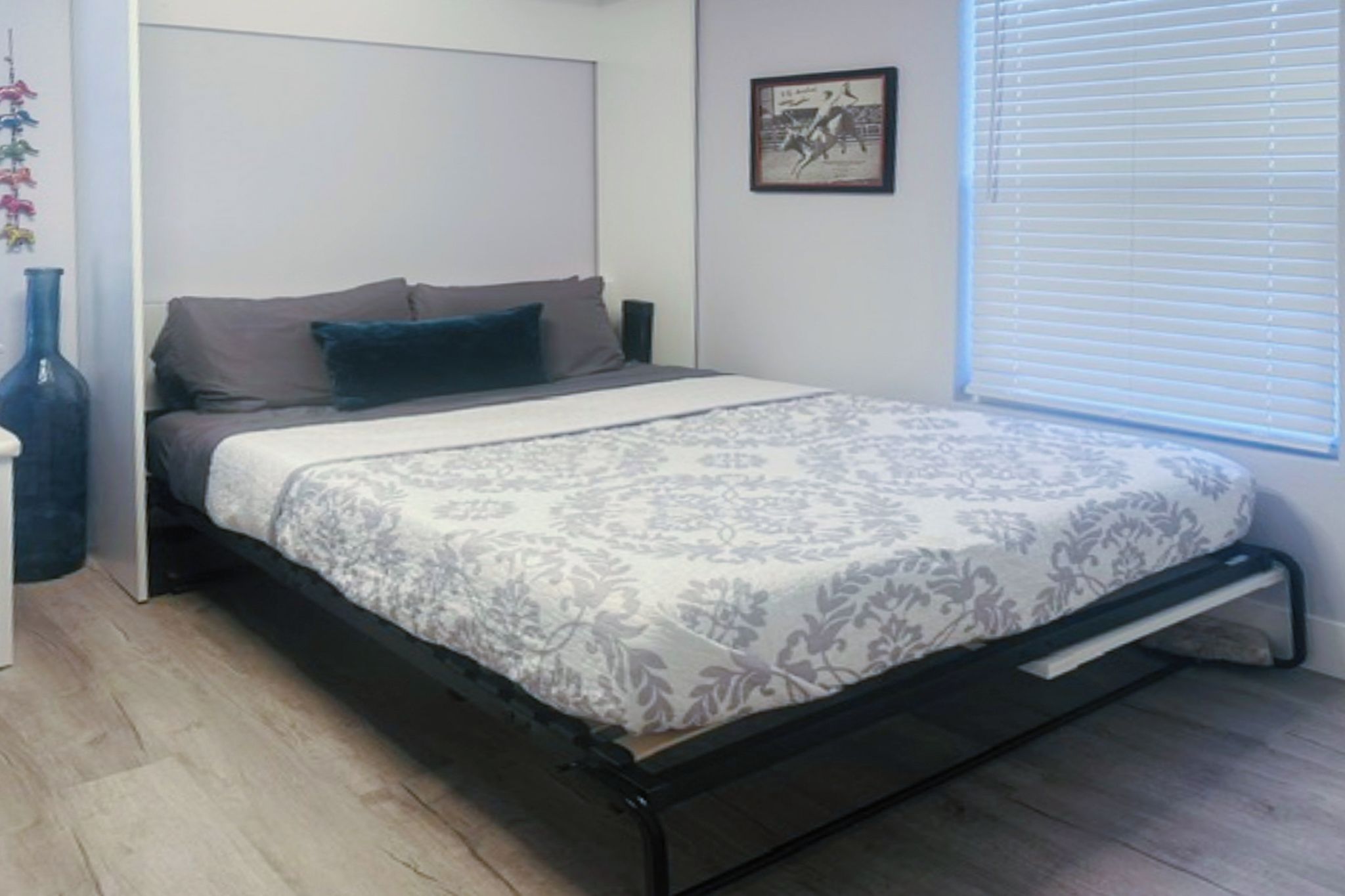 A bedroom with a bed, a nightstand, a wall-mounted picture, and a window with blinds.