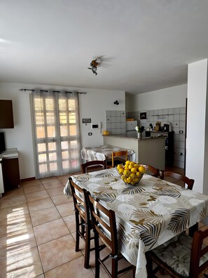 Ein Essbereich mit Tisch für vier Personen, eine Küche im Hintergrund und ein Fenster mit Vorhängen.