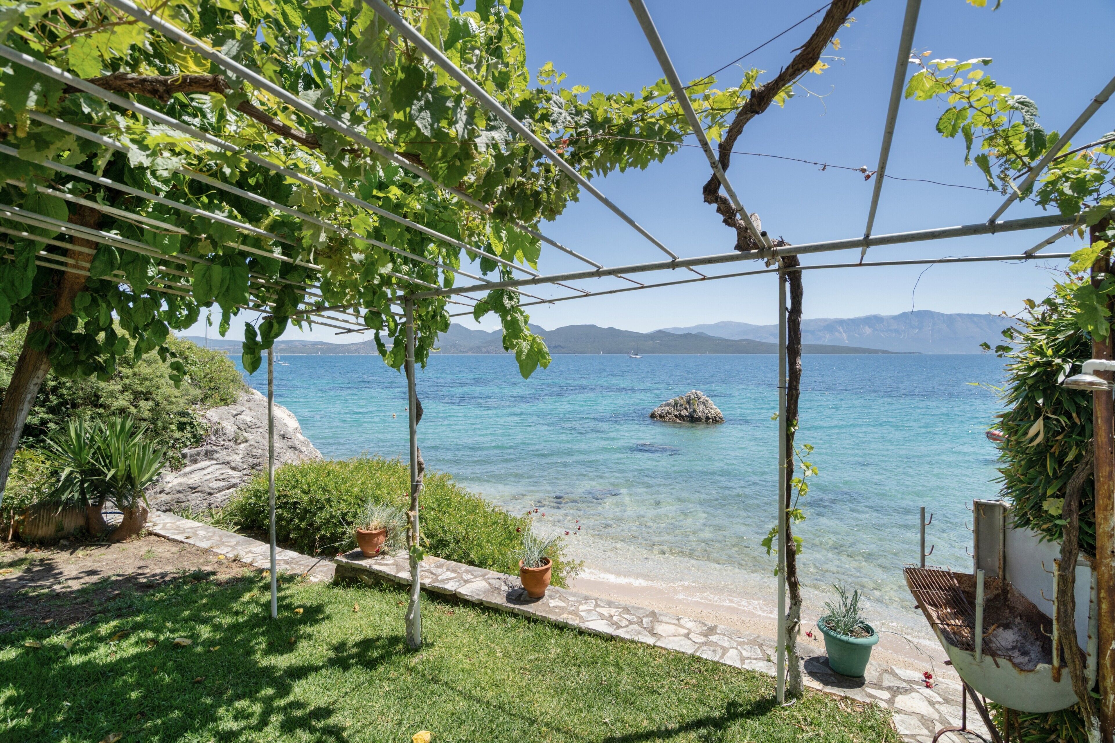 Ein Garten mit einer Pergola, Topfpflanzen und freiem Blick auf ein Gewässer und Berge.