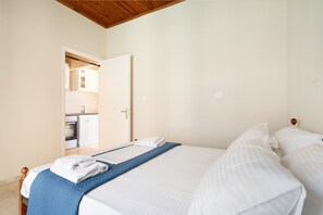 A bedroom with a bed, a blue bed runner, white bedding, a wooden ceiling, and a door leading to a kitchen. - Secret Beach A Apartment with Sea View, Wi-Fi, and Air Conditioning (Nikiana)