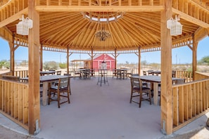 A wooden gazebo with tables and chairs, a red barn in the background, and a clear sky.