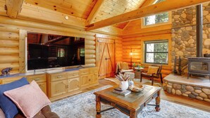 A log cabin interior with a stone wall, wooden furniture, a fireplace, and a large TV. - Forest, Fairways, Lake Almanor Days! Lake Love (Westwood)