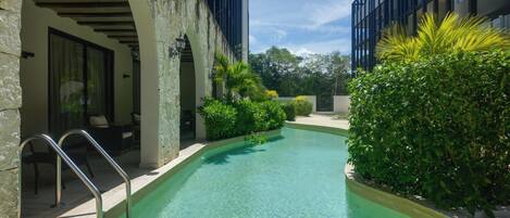 A swimming pool with a stone wall and metal railings, surrounded by greenery and modern buildings.
