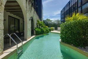 A swimming pool with a stone wall and metal railings, surrounded by greenery and modern buildings. - Resort Living in Tulum! Walkable Downtown Condo (Tulum)