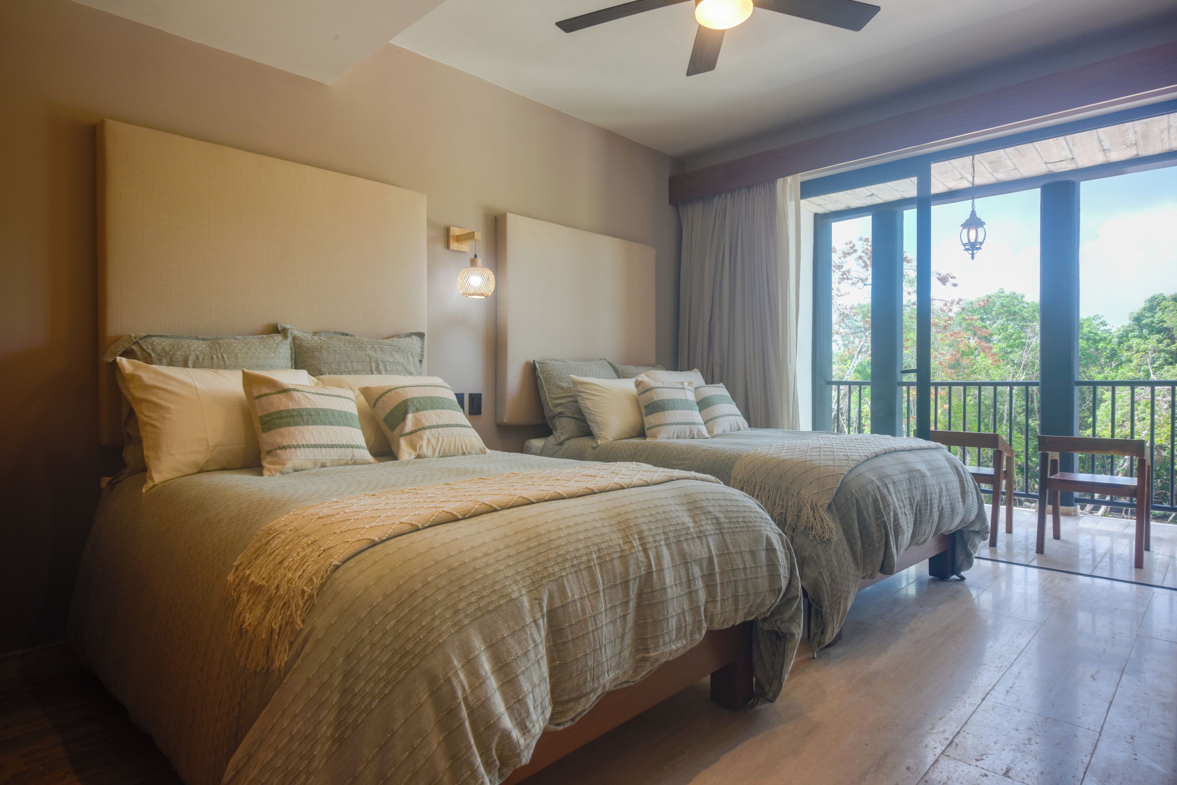 A hotel room with two beds, a ceiling fan, a nightstand, and a balcony with a view of trees.