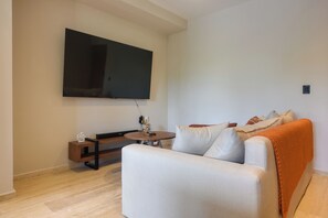 A living room with a flat-screen TV mounted on the wall, a round wooden coffee table, a white sofa, and a patterned throw blanket. - Resort Living in Tulum! Walkable Downtown Condo (Tulum)