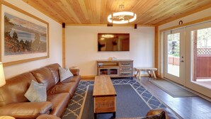 Techo de madera, un sofá de cuero marrón, una mesa de centro de madera, un televisor y un gran ventanal corredizo.
