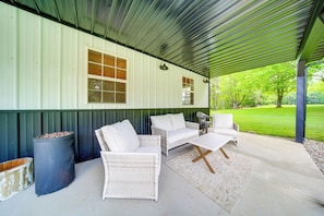 A covered patio with wicker furniture, a trash can, and a window overlooking a green lawn.