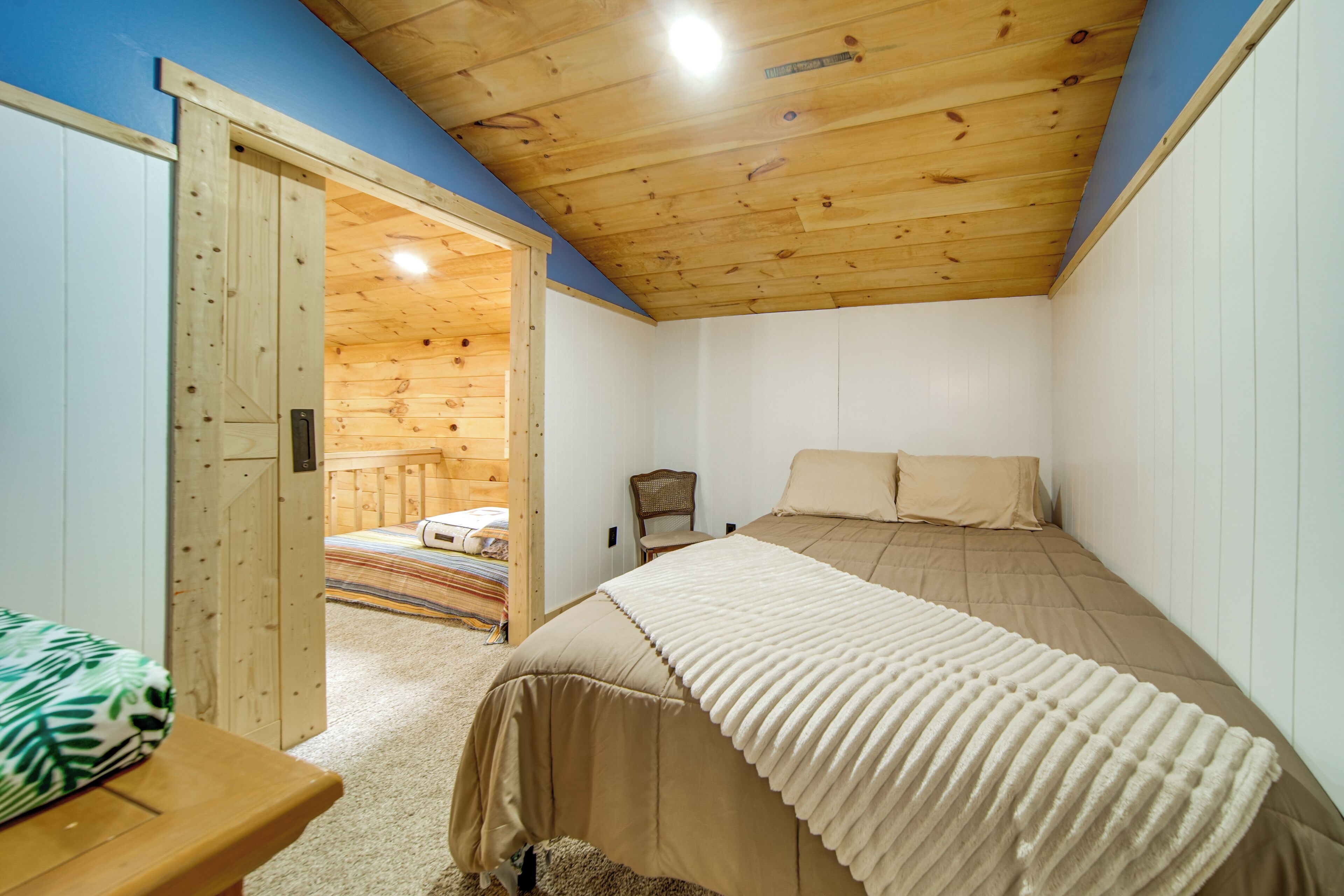 A bedroom with a wooden ceiling, a bed with a beige bedspread, a chair, and a wooden door leading to another room.