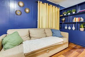 A beige sofa with a white throw blanket, a green pillow, and a blue pillow. There are decorative wall sconces and a curtain. A blue bookshelf with plants and books is visible. - 1 Mi to UT & Med Centers! Updated Austin Studio (Austin)