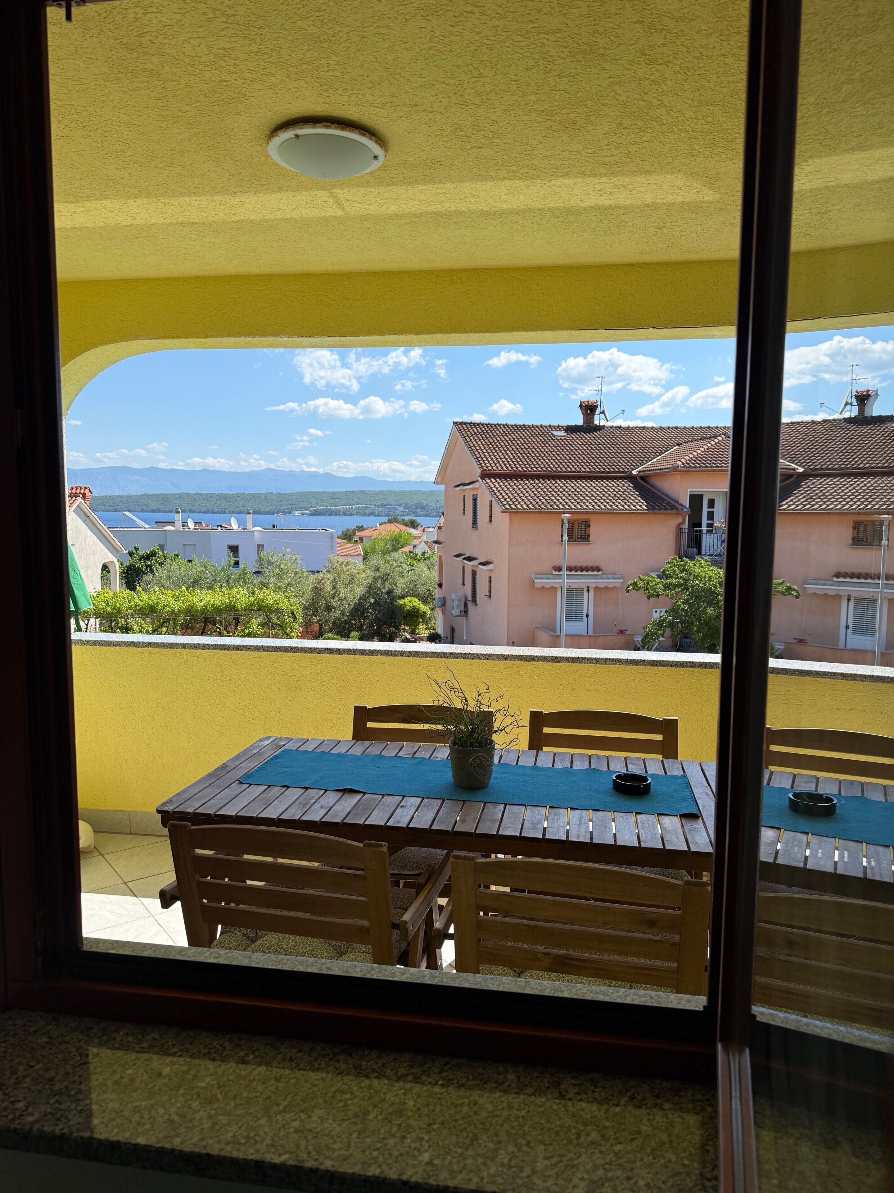 Ein Balkon mit Tisch und Stühlen, einem Blumentopf und Blick auf Häuser und Berge.