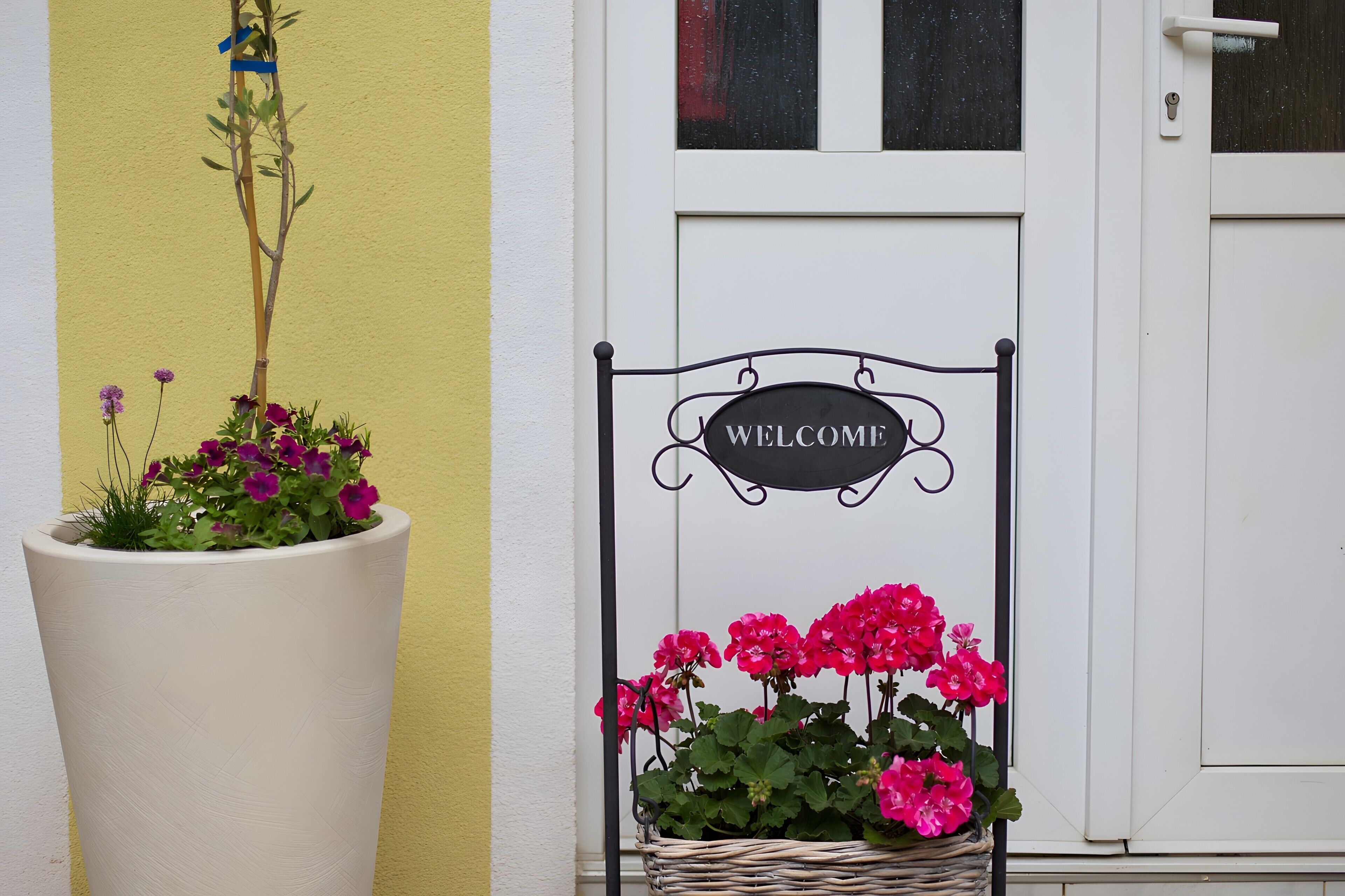 Una puerta blanca con un letrero que dice 'WELCOME', una maceta grande con flores y una cesta de mimbre con flores.