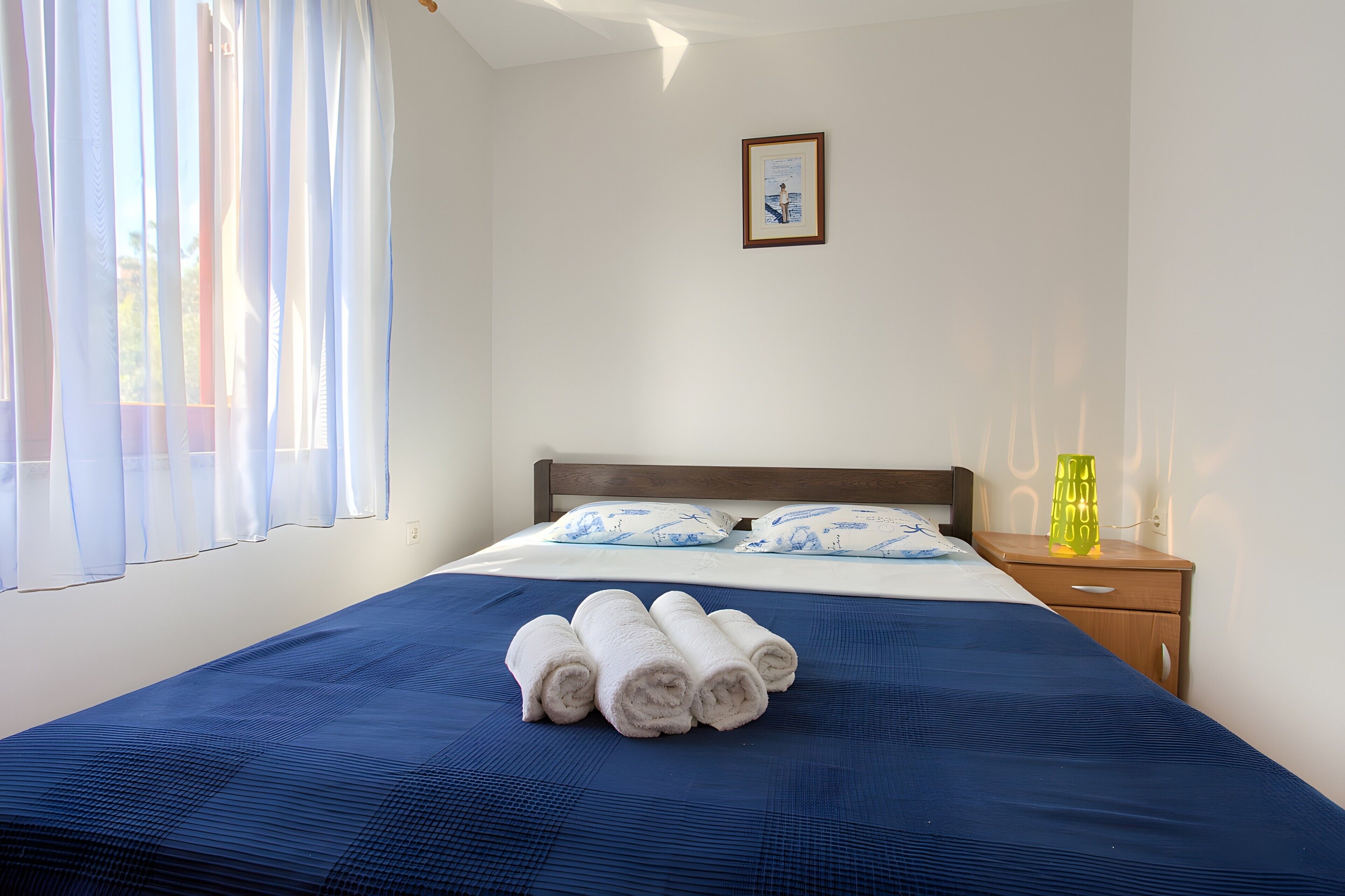 A neatly made bed with rolled towels, a bedside table with a lamp, and a framed picture on the wall.