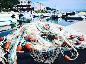 Uma marina com barcos atracados, um monte de redes de pesca e edifícios ao fundo.