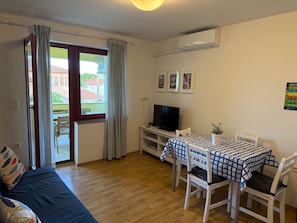 Quarto com piso de madeira, sofá, mesa de jantar com cadeiras, televisão e janela com cortinas.