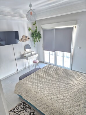 Ein Schlafzimmer mit Bett, Schreibtisch, Stuhl, einer hängenden Pflanze und einem Fenster mit Jalousien.