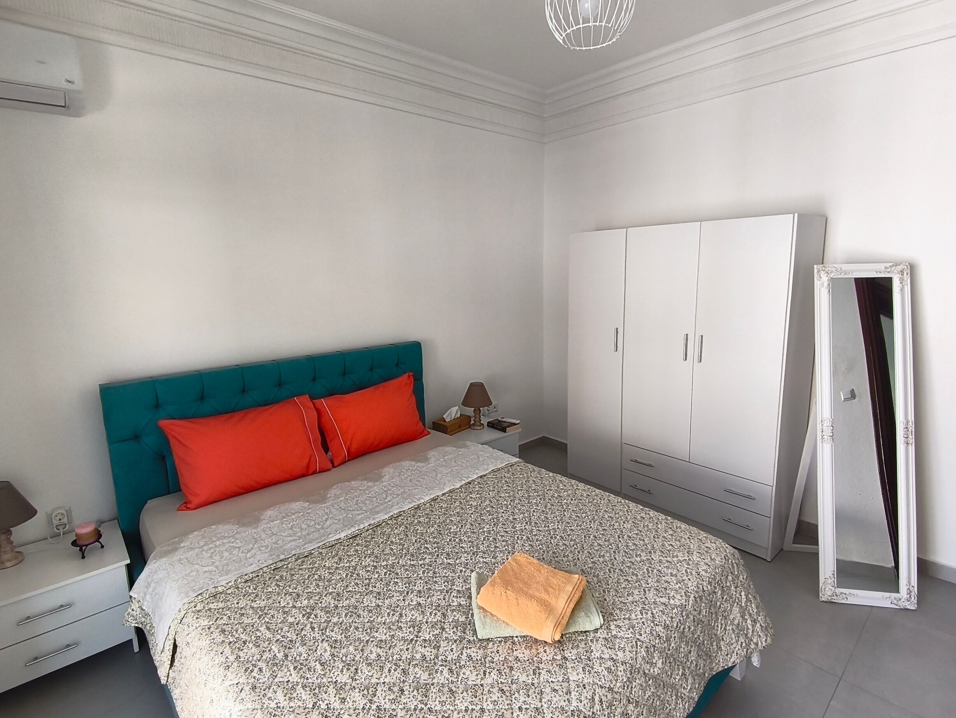 Une chambre à coucher avec une tête de lit de couleur turquoise, une armoire blanche, un miroir et un lit recouvert d’une couvre-lit à motifs.