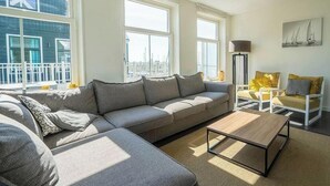 A modern living room with a sectional sofa, a coffee table, and a chair. There are large windows providing a view of the city. - Holiday Home in Uitdam With Terrace (Uitdam)