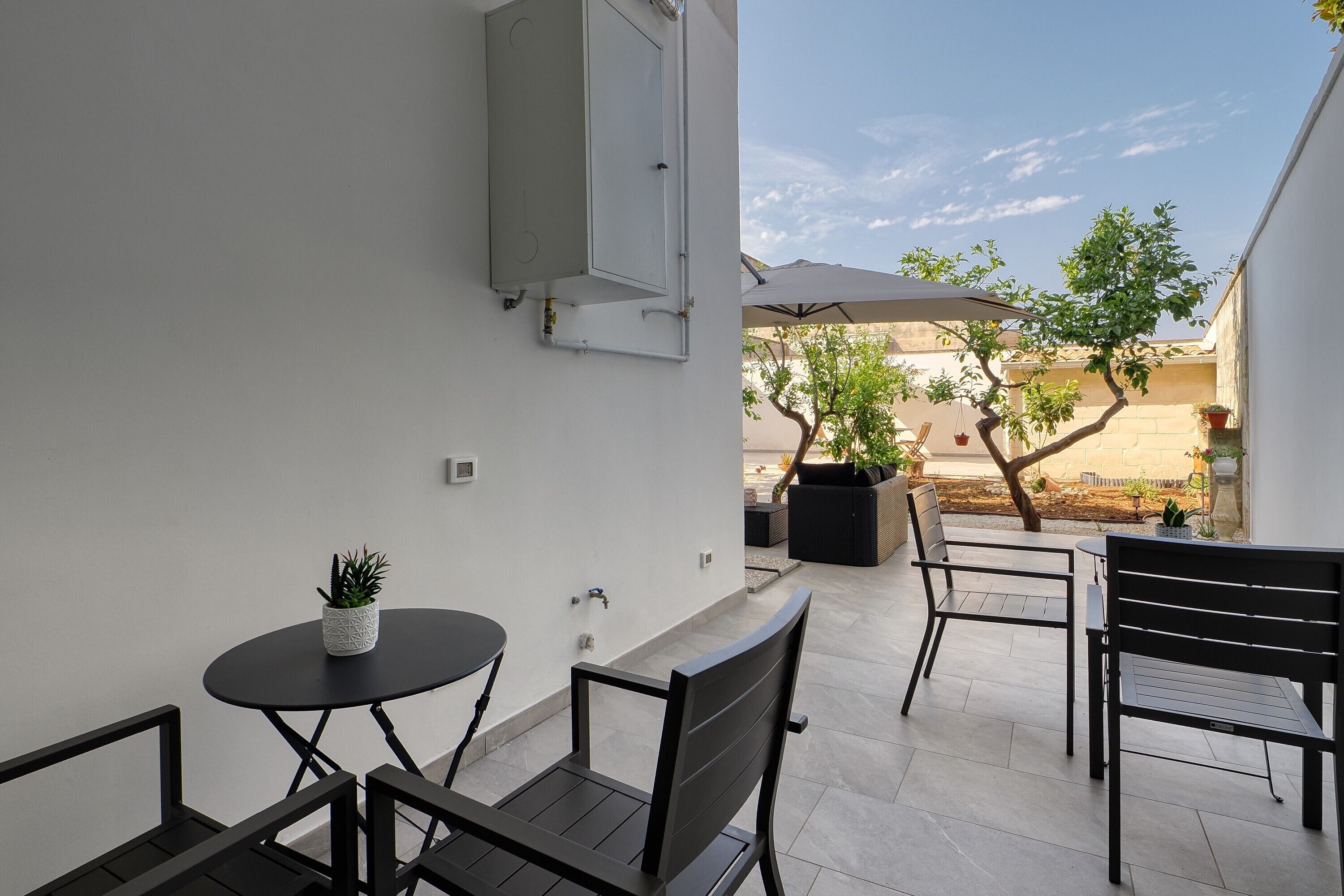Eine Terrasse mit rundem Tisch und zwei Stühlen, einem Blumentopf und einer weißen Wand mit einem Stromkasten.
