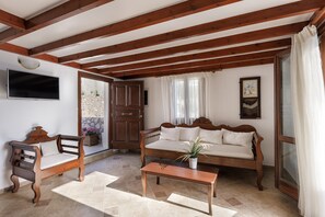 Ein Wohnzimmer mit einer Holzbänke, einer Couch und einem Couchtisch, einem an der Wand montierten Fernseher und einer sichtbaren Steinwand durch eine Tür hindurch.