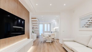 A modern living room with a flat-screen TV, a white sofa, a dining area with a round table and chairs, and a kitchen in the background. - Cagliari Suite Monti Fly – Renovated Central Apartment Near San Benedetto Market (Cagliari)