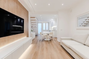 Ein modernes Wohnzimmer mit einem Flachbildfernseher, einem weißen Sofa, einem Essbereich mit rundem Tisch und Stühlen sowie einer Küche im Hintergrund.