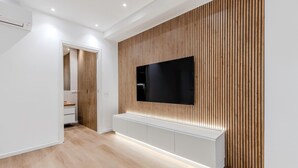 A modern living room with a flat-screen TV mounted on a wooden panel wall, a white entertainment unit below, and a hallway leading to a bathroom. - Cagliari Suite Monti Fly – Renovated Central Apartment Near San Benedetto Market (Cagliari)