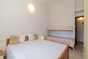Una stanza con un letto singolo, un letto a castello, un comodino, una lampada a parete e un'uscita che dà su un'altra stanza.