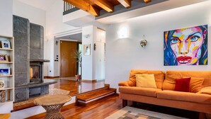 Uma sala de estar com lareira, um sofá, um quadro colorido e uma escada que dá para outro cômodo.