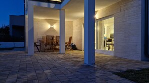 Un espacio al aire libre con patio, un comedor y una silla plegable, una puerta de vidrio que da a una cocina y una pared exterior blanca.