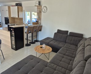 Una sala de estar moderna con un sofá seccional gris, una mesa de centro de madera y una zona de cocina con refrigerador, horno y taburetes de bar.