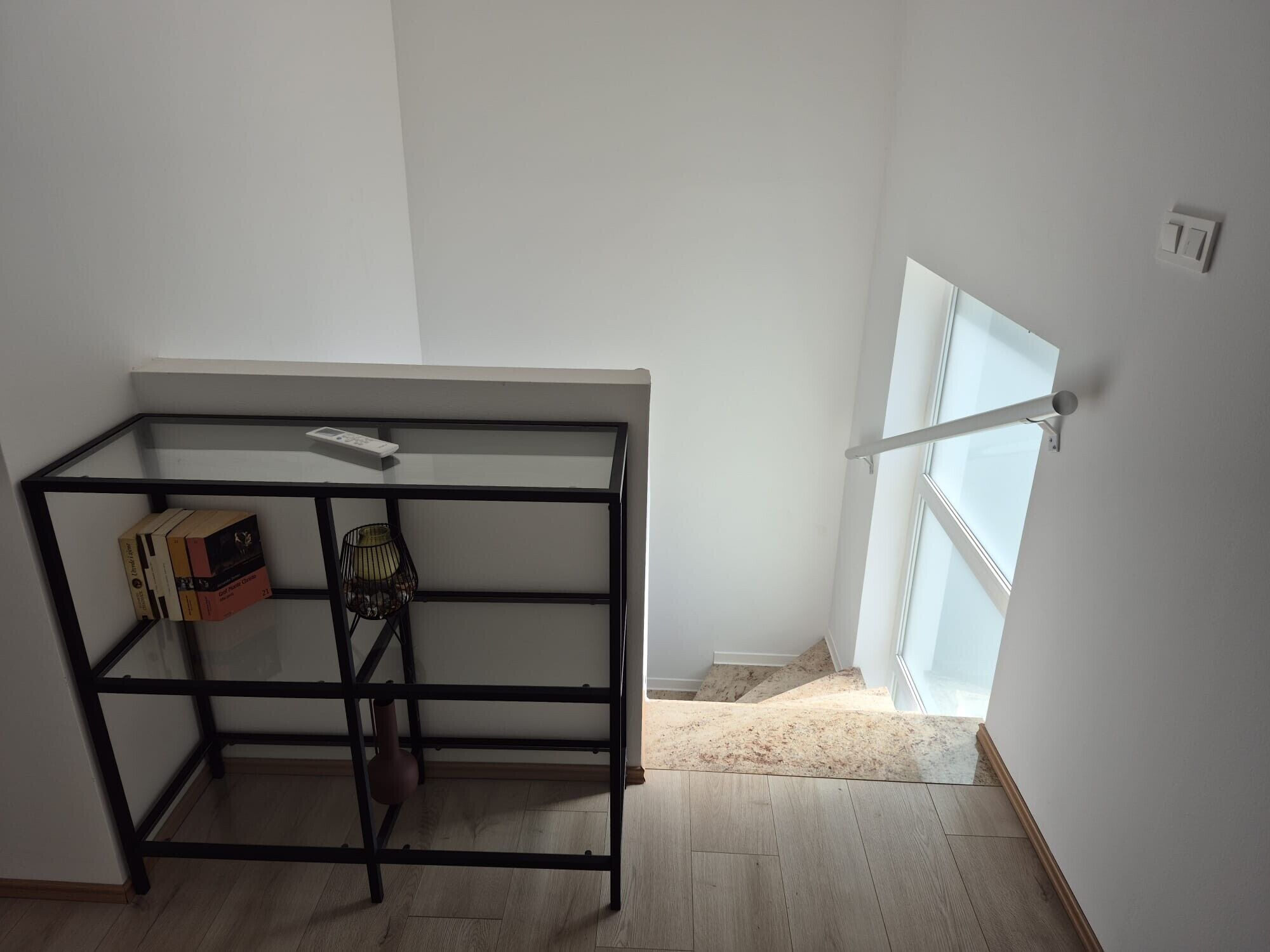 Een kamer met een zwarte metalen schap, een klein tafeltje met een afstandsbediening, en een raam met uitzicht op een trap.