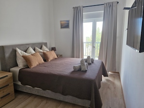 Un dormitorio con cama, almohadas, una mesita de noche, televisión y ventana con cortinas.