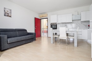 Ein modernes Wohnzimmer mit einer grauen Couch, einem Esstisch mit Stühlen, einer Küche mit weißen Schränken und einer roten Tür.