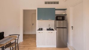 Eine kompakte Küche mit Mikrowelle, Kühlschrank und einer kleinen Spüle. Im angrenzenden Raum befindet sich ein Esstisch mit Stühlen sowie ein Bücherregal.