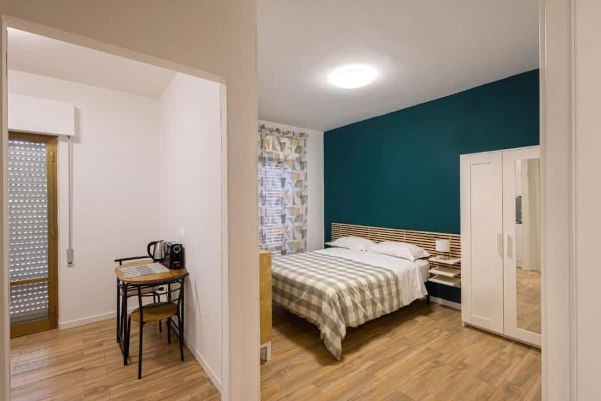 A bedroom with a bed, a bedside table, a chair, a wardrobe, and a door leading to another room.