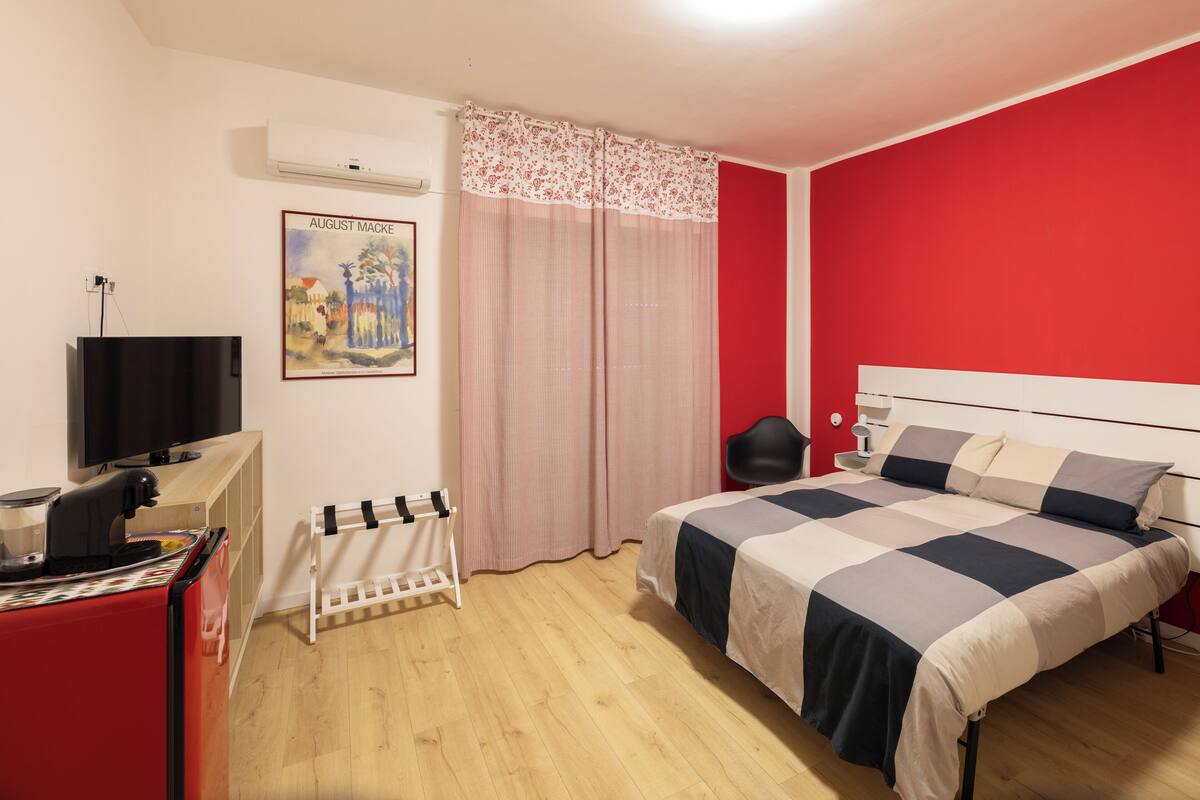 A bedroom with a bed, a TV, a red wall, and a painting.