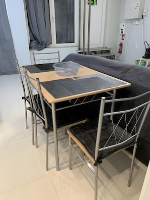 Ein ausklappbarer Tisch mit einer transparenten Schüssel, graue gepolsterte Stühle, ein Fenster mit Vorhängen und ein Feuerlöscher an der Wand.