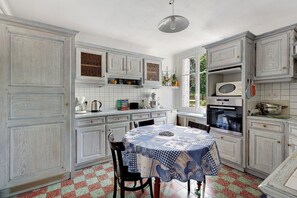 Une cuisine avec des meubles blancs, une table recouverte d’une nappe bleue et une fenêtre donnant sur un beau paysage.